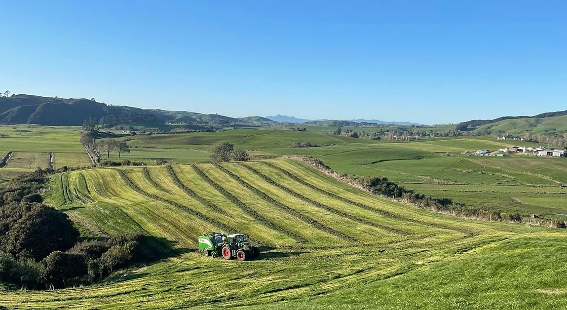 Aerial view of a tractor and baler working a large mown paddock across the rolling Waikato hill country