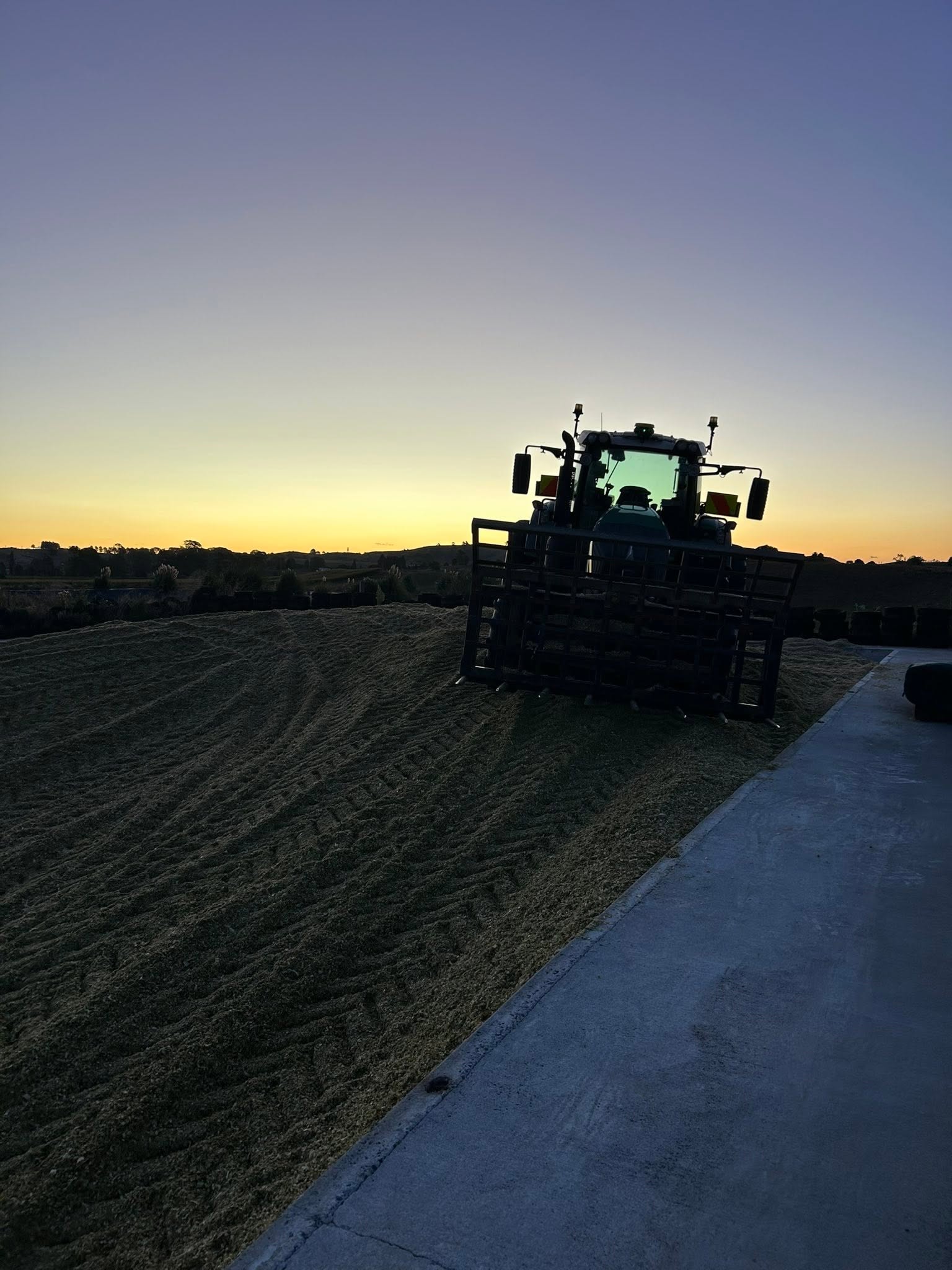 Tractor silhouetted on top of a maize silage stack against an orange and purple dusk sky