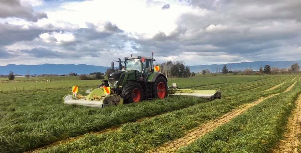Fendt tractor mowing a grass silage paddock with front and rear mower-conditioners, hill ranges under a cloudy sky behind