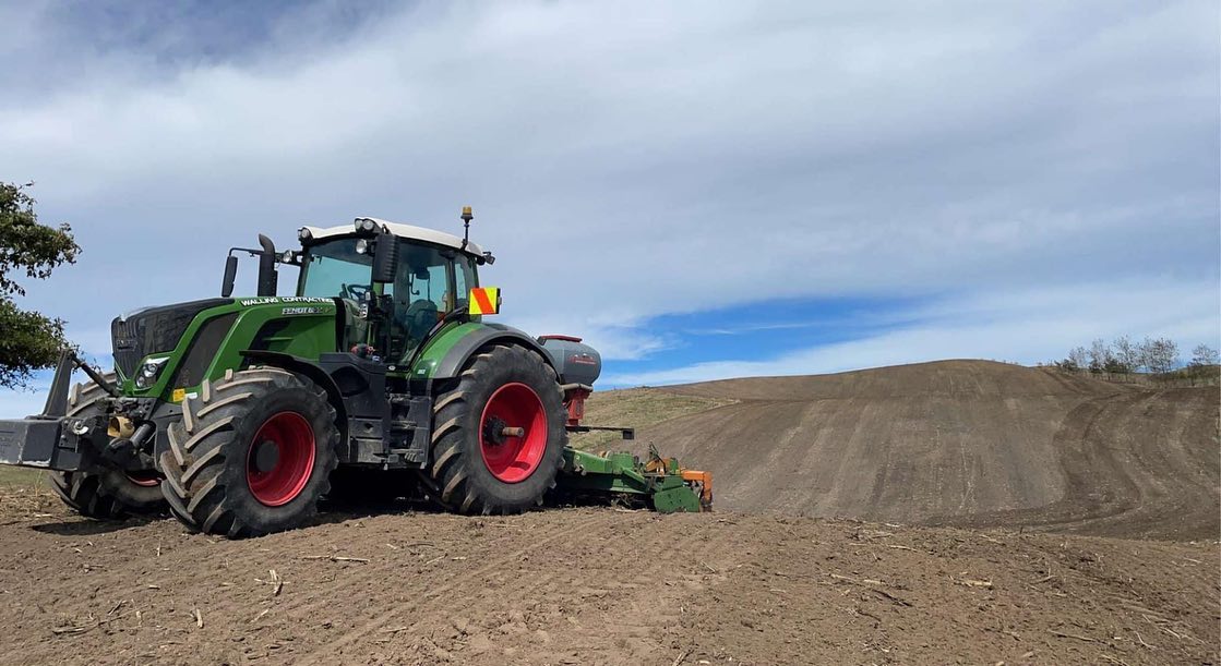 Green Fendt tractor in Walling Contracting livery pulling a power harrow and seed drill combination across freshly cultivated brown ground on a hill paddock under a bright sky