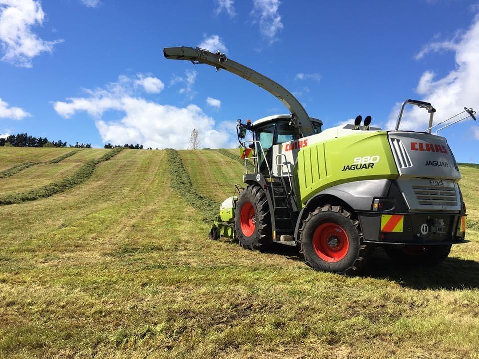 Fendt tractor towing an orange trailed sprayer with the boom extended, working across a paddock of long grass under an overcast sky with ranges on the horizon